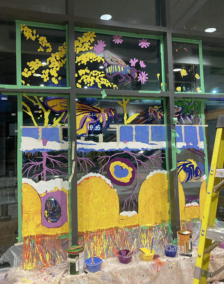 An image of a partially completed painting on a downtown window showing bees hovering over a landscape. 
