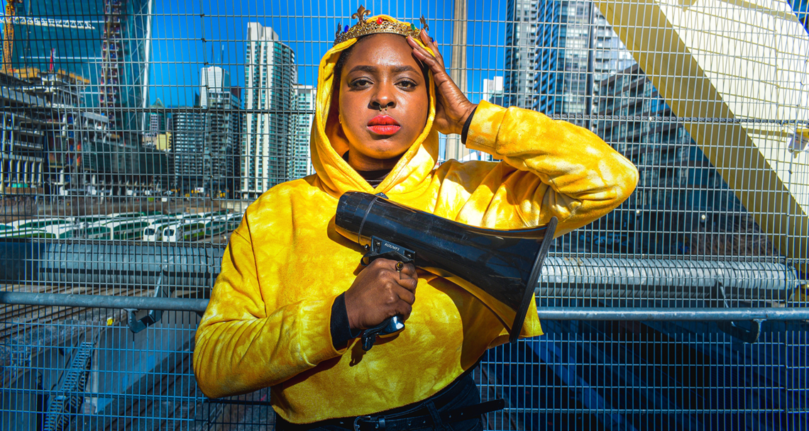 A Black woman wearing a crown and bright yellow hoodie stands against a wire fence. One hand is on her head, and the other holds a megaphone