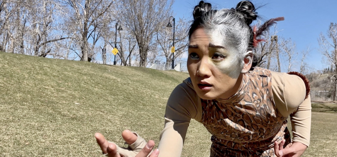 A dancer with silver paint on half of her face gestures in an empty field