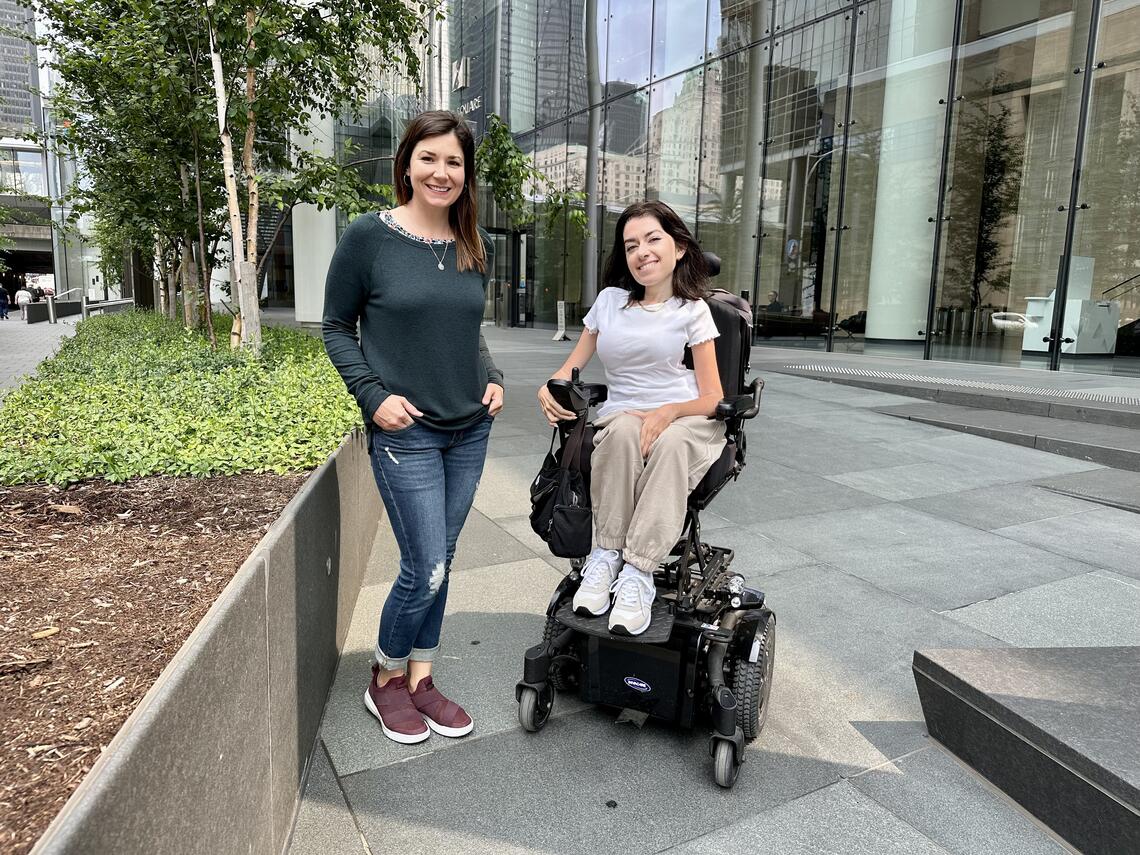 A woman in a wheelchair is positioned next to a woman standing and both are smiling at the camera
