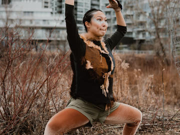 A dancer stands in a wide squat, hands up in the air. Downtown Calgary is visible behind them.