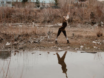 A dancer stands and arches their back, arm raised. Their reflection is visible in a pond in front of them.