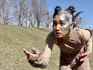 A dancer with silver paint over part of her face gestures in an empty field