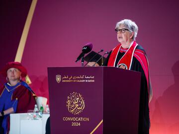 A woman in convocation regalia stands behind a podium giving a speech