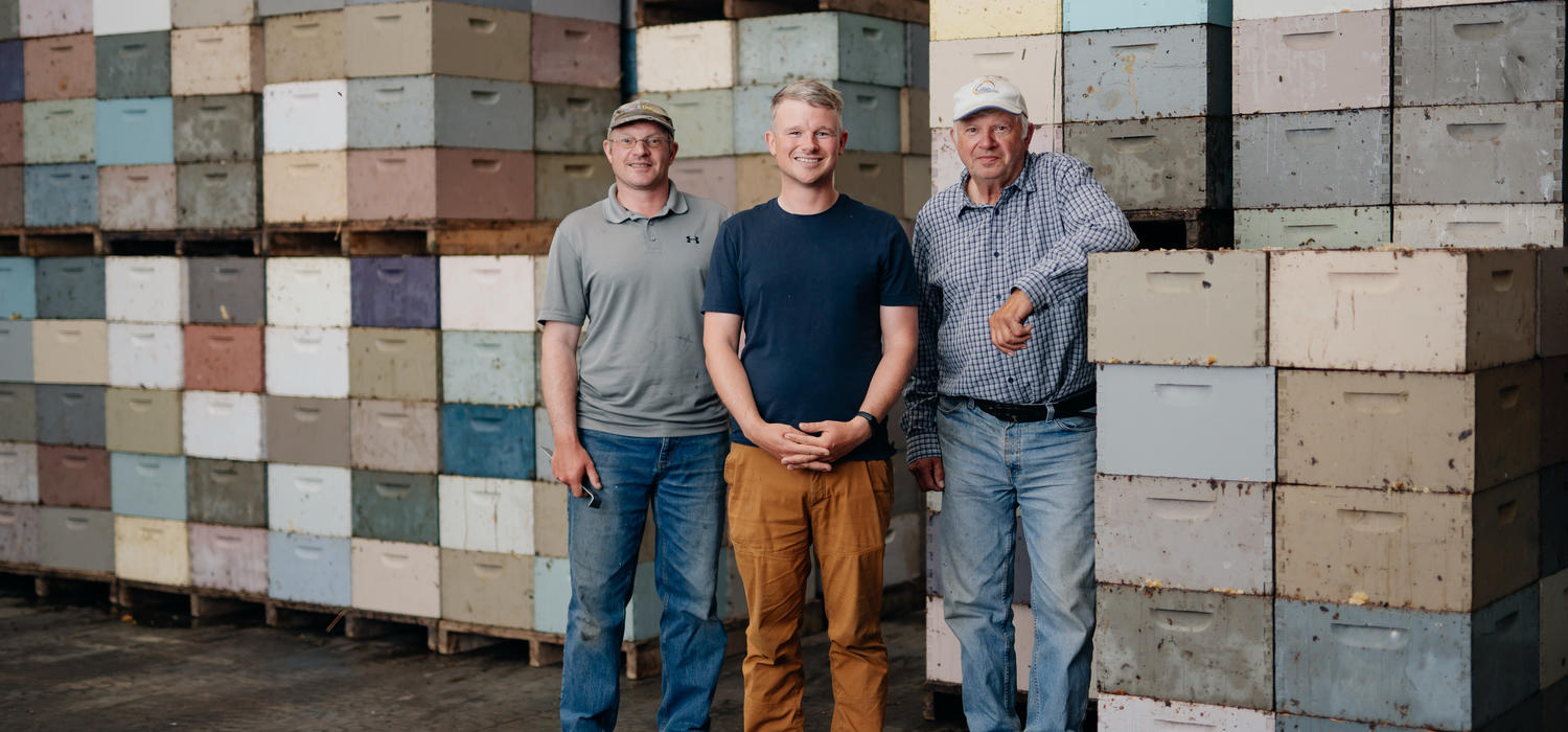 Three men stand in front of palettes of honey