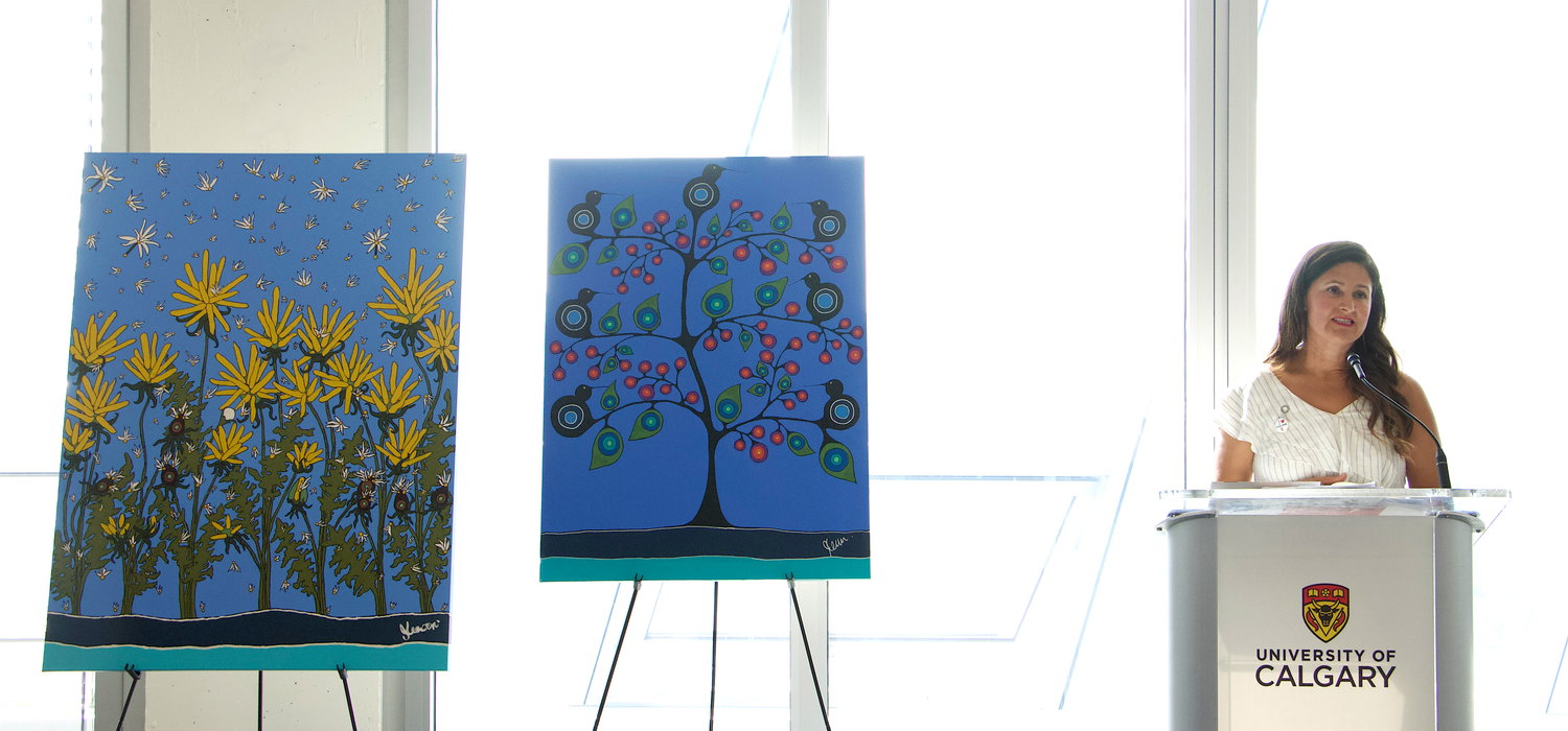 Dr. Jennifer Leason talks at a podium with two of her colourful paintings off to the side.