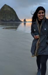 Alumna Tonie Minhas stands on a beach at sunset. She is wearing a jacket, holding a cup of coffee, and smiling at the camera. The sky is grey and gold behind her.