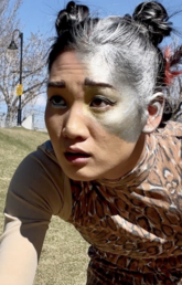 A dancer with silver paint on half of her face gestures in an empty field