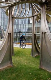 A close up shot through a statue on UCalgary campus shows a cyclist and a building in the distance