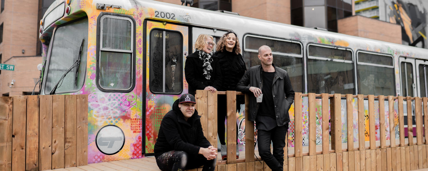 Individuals sit and stand in front of a colourfully painted LRT car in downtown Calgary