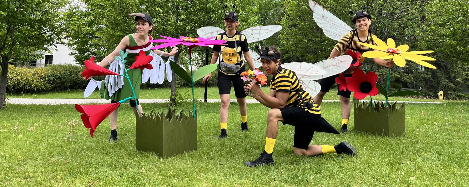 Student performers in Wagonstage Theatre in an action shot. They're wearing bee or bird costumes and are outside on the campus lawn. Brightly coloured paper flowers are placed beside and behind them.