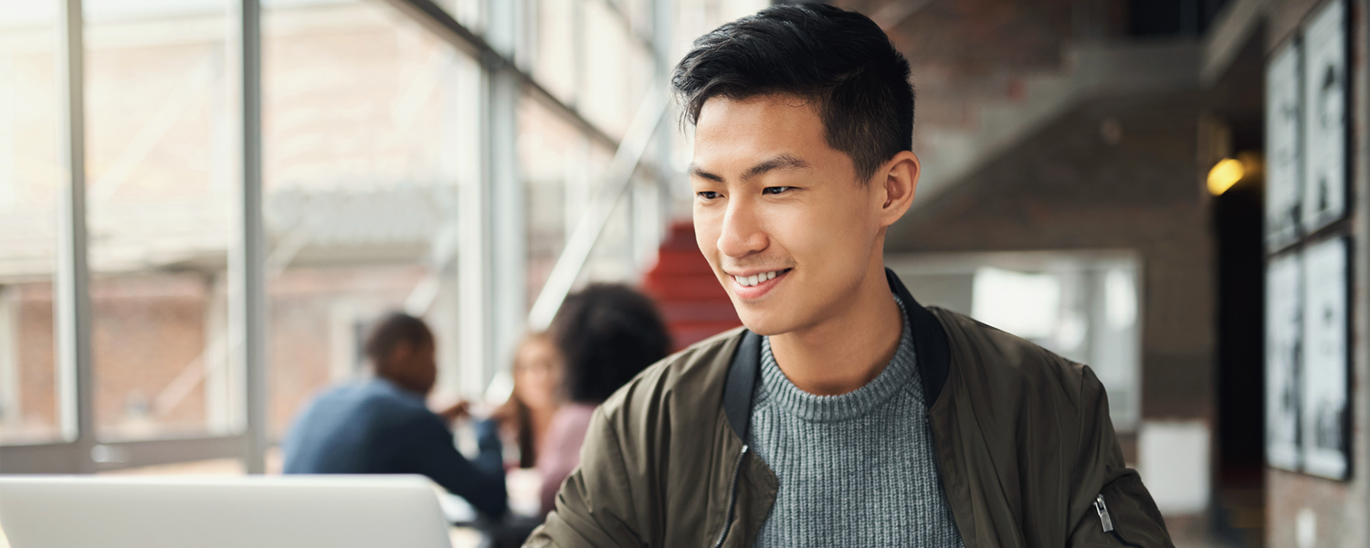 Student smiles has he studies at his laptop. Blurred students are visible in the background.