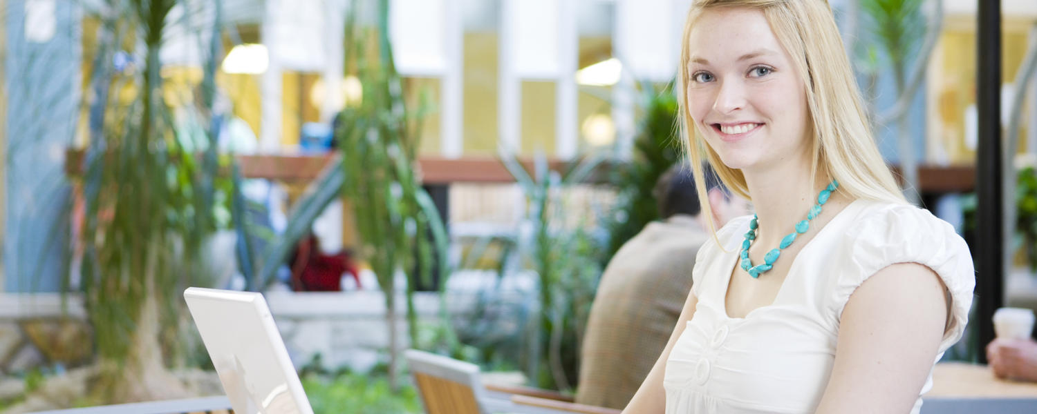 A student smiles at the camera. She is working on a laptop in one of the student spaces on campus.