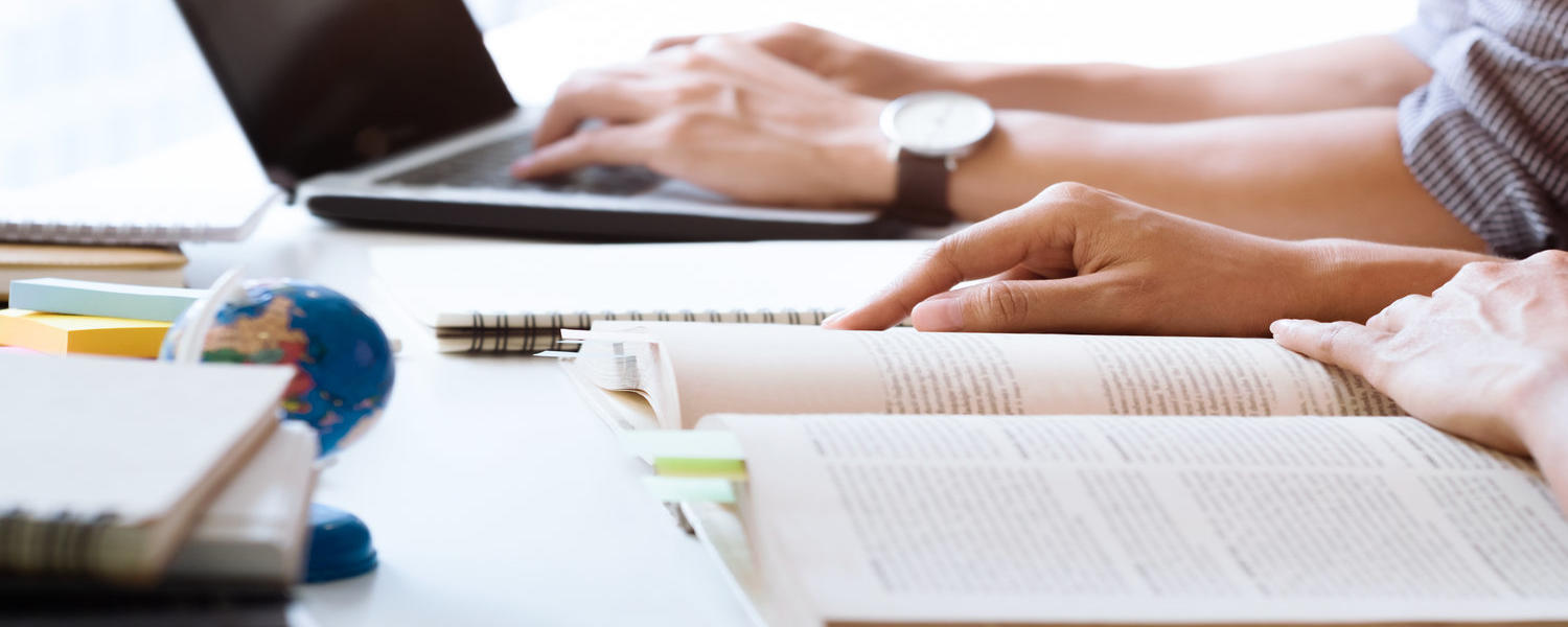 stock photo: students study together with a book and a laptop. Hands and desk appear in picture.