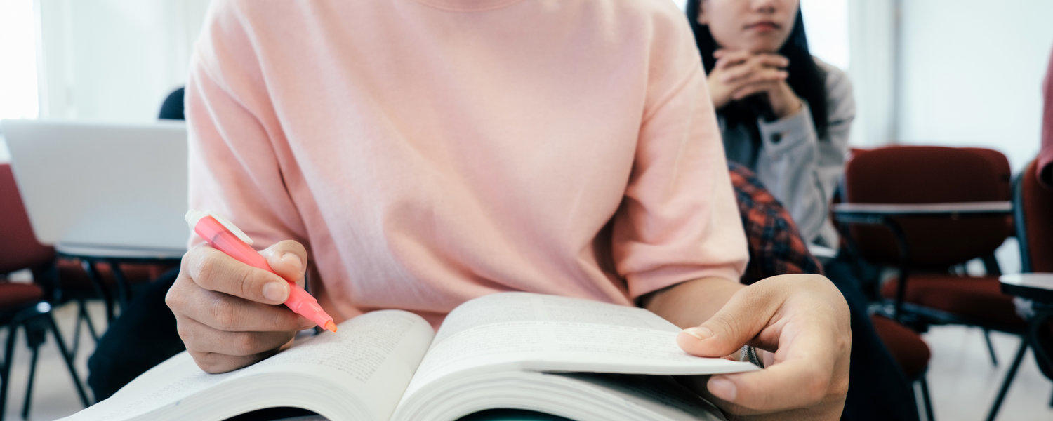 Stock photo: a student highlights a passage she is reading in a book. Others are visible seated at desks behind her. Classroom setting.