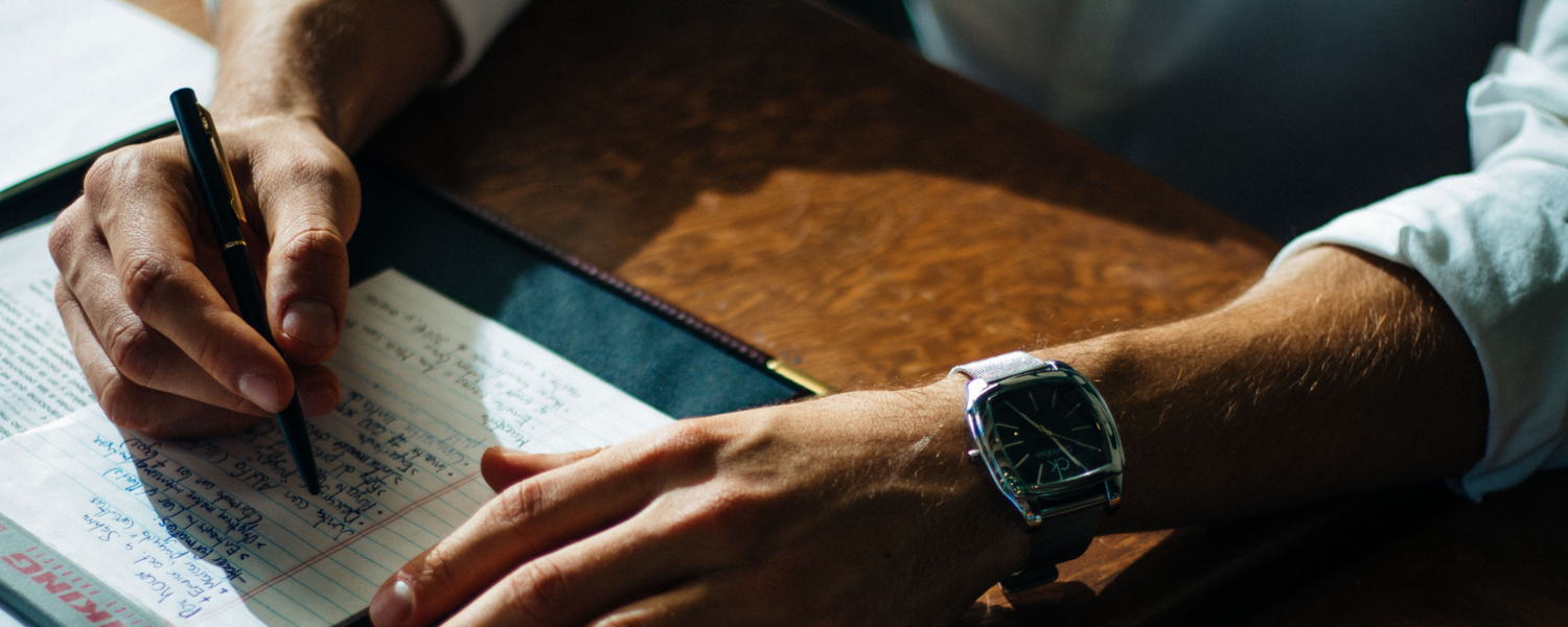 Photo of a person at a desk, writing with a pen on a notepad. Only the person's arms and hands are visible. They wear a heavy watch.