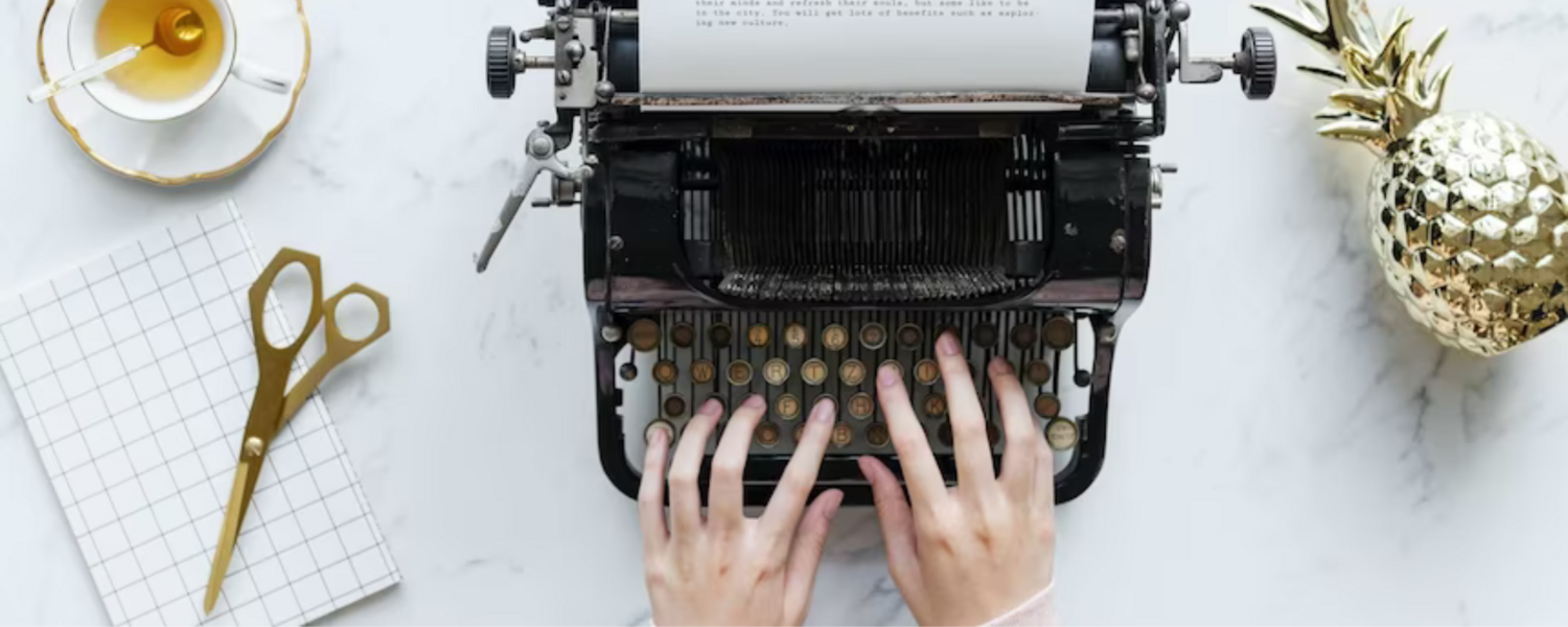 Photo of hands at typewriter, a cup of tea and pair of scissors to the left, and a golden pineapple inexplicably at the right.