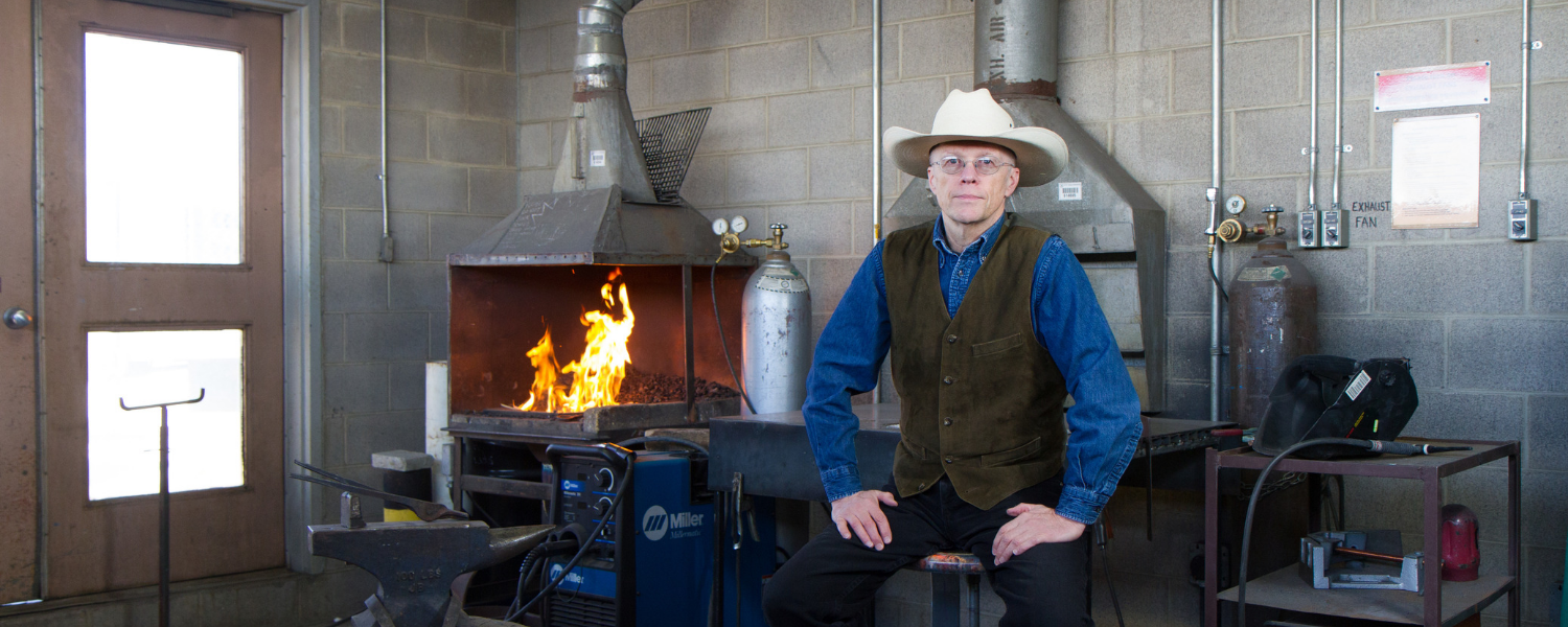 Man in a cowboy hat sits in a blacksmith workshop, fire burning behind him.
