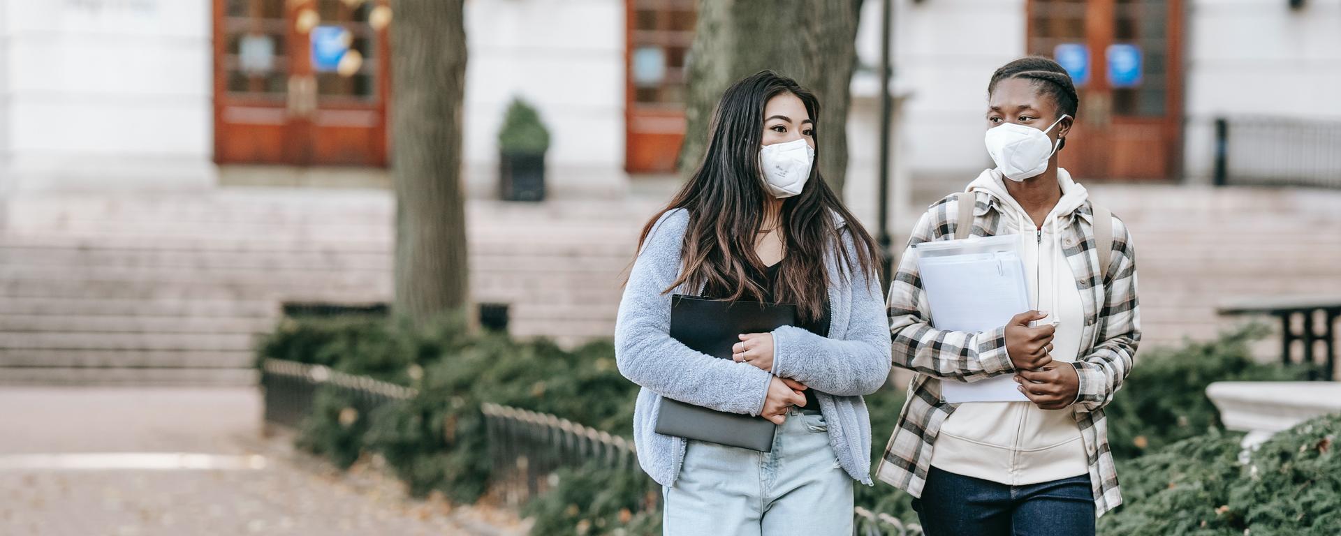 Two young women walk on a campus caryring books and wearing masks