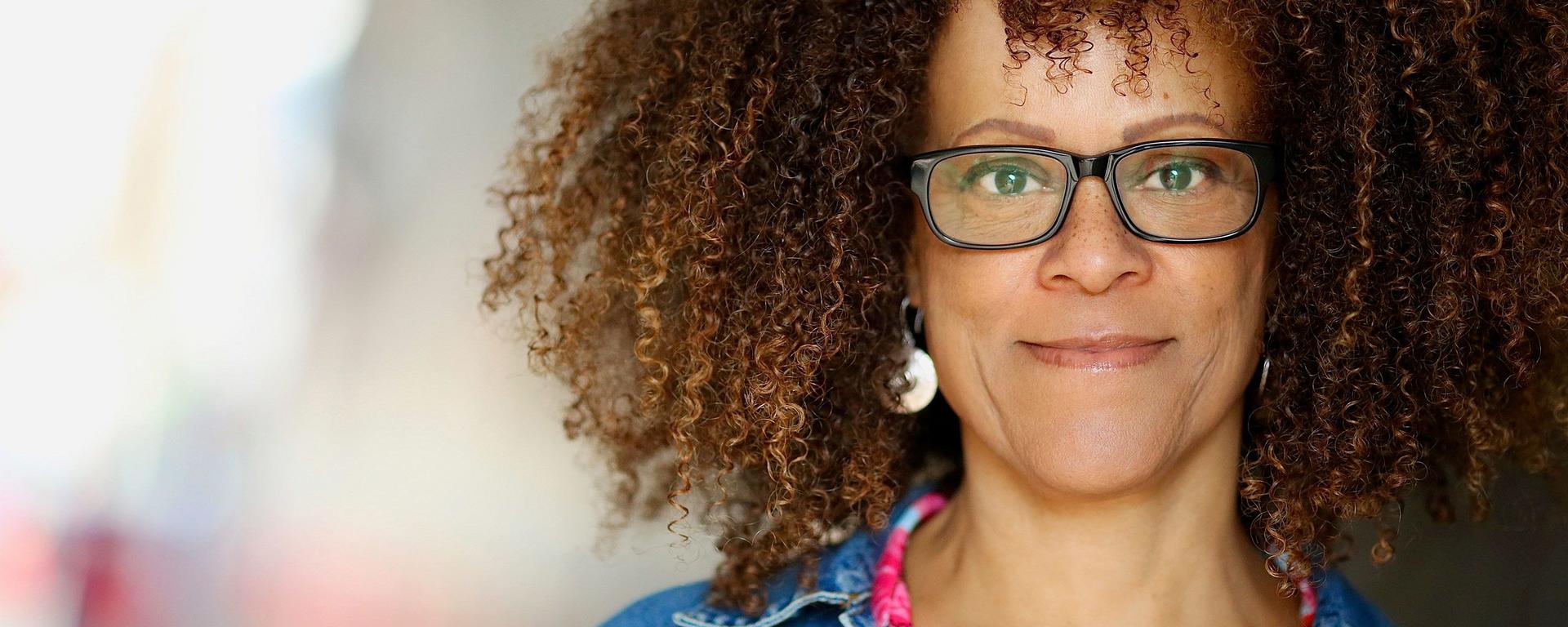 Head shot of 2021-22 Distinguished Visiting Writer Bernardine Evaristo. She wears silvery earrings, dark rimmed glasses, and a jean jacket over a pink floral blouse.