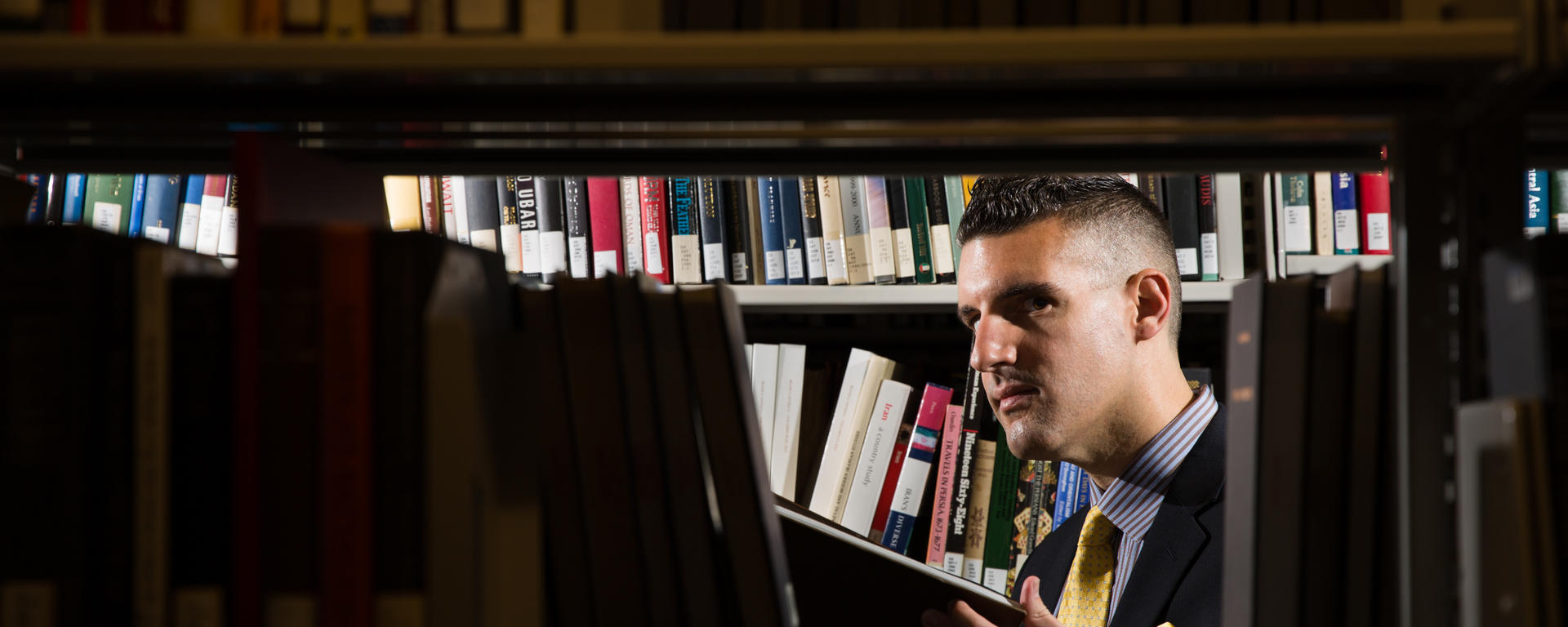 PhD in English Literature, Dr. Anthony Camara is seen through the stacks of books in the library
