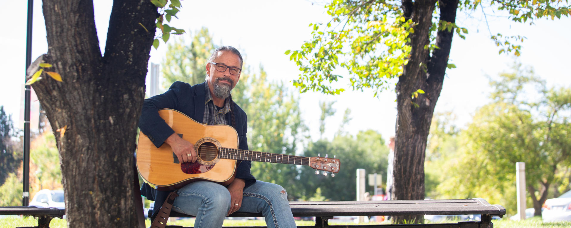 Classics and Religion Senior Instructor Craig Ginn sits outside on a picnic bench with his guitar