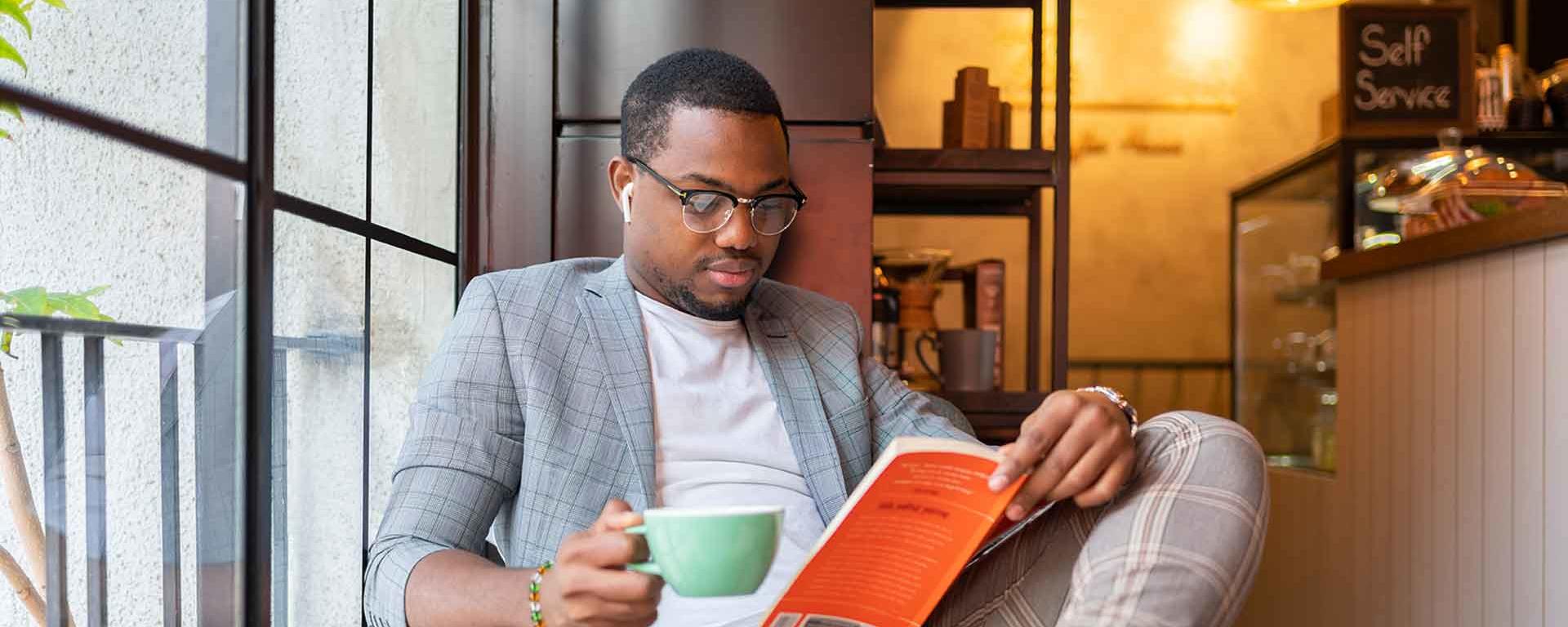 A Young Man Reading Book And Listening Music On Smartphone At Coffee Shop
