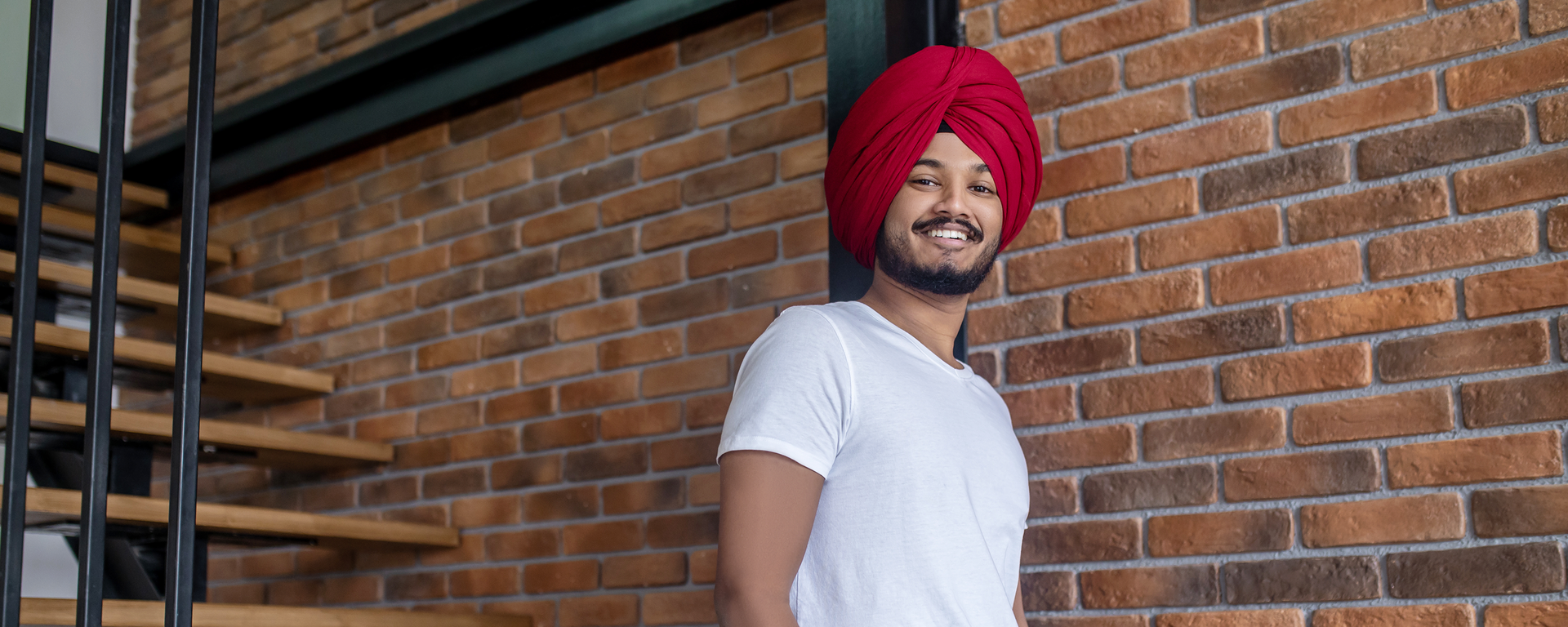 A young Sikh man walks down stairs carrying a laptop. He wears a turban and he is looking towards the camera and smiling.