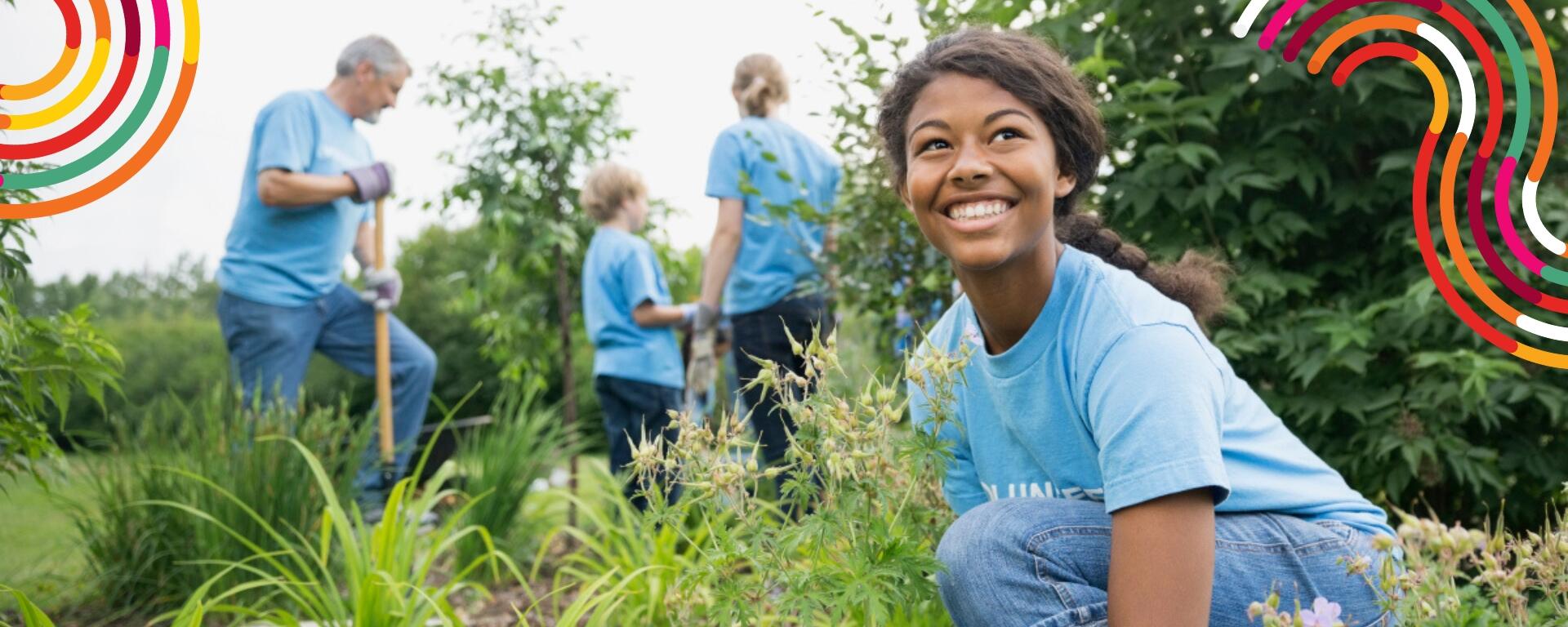 A young person in gardening gloves smiling, with a group of three other people in the background. They are working outdoors. 