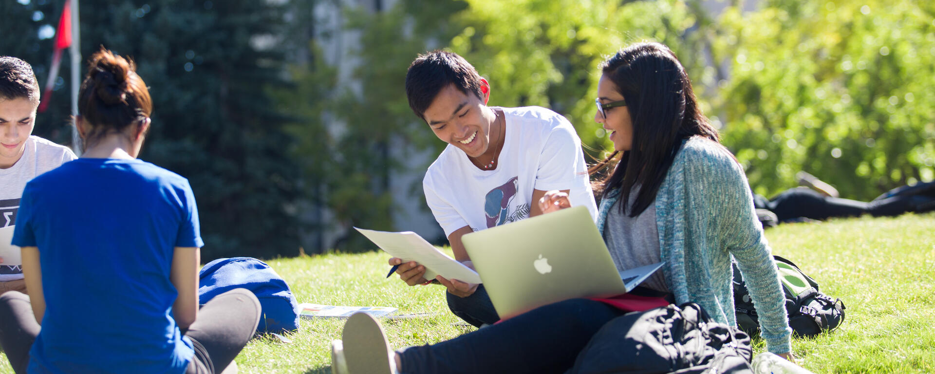 Students sit outside together, looking at papers and a laptop