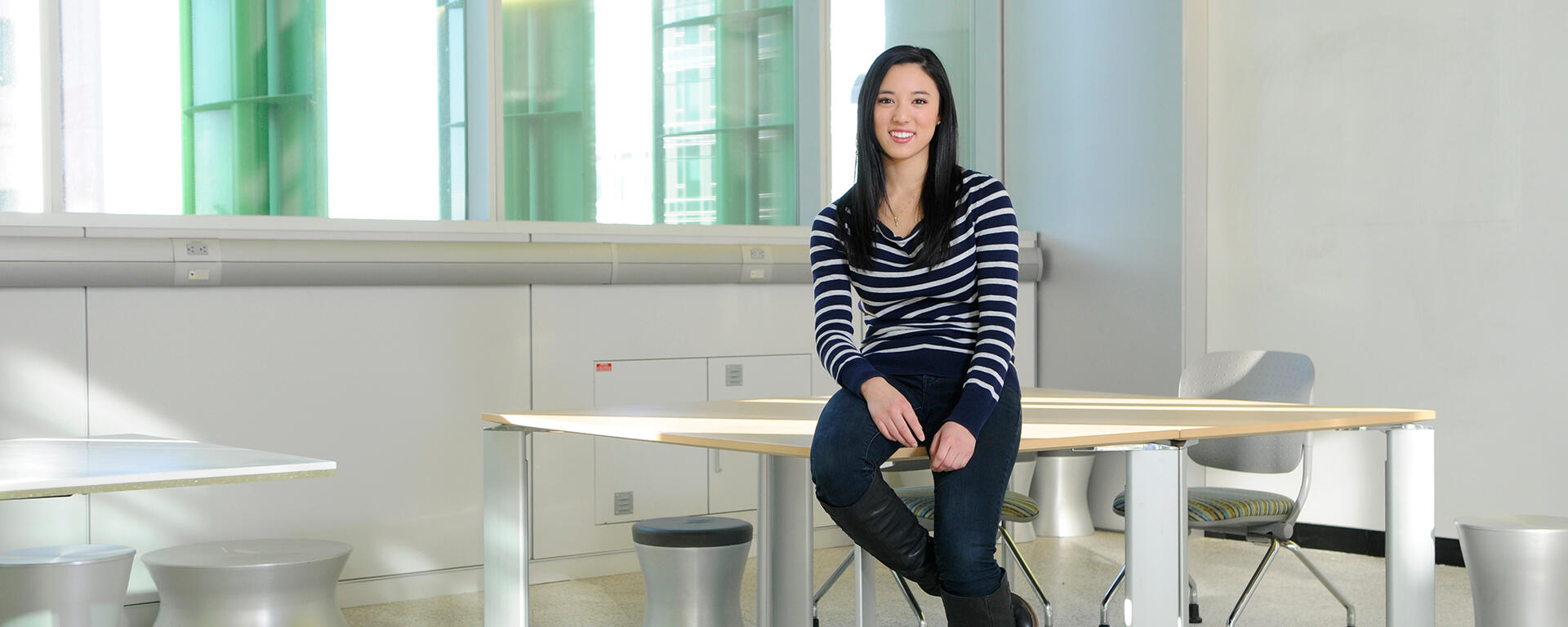 A person perches on a desk in an empty classrom. They are smiling.