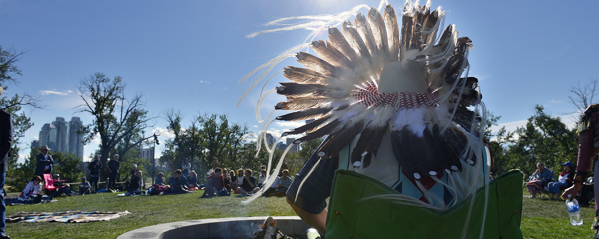 A man in a headdress is visible from the back, at Campfire Chats. People are seated on the ground in front of him listening.