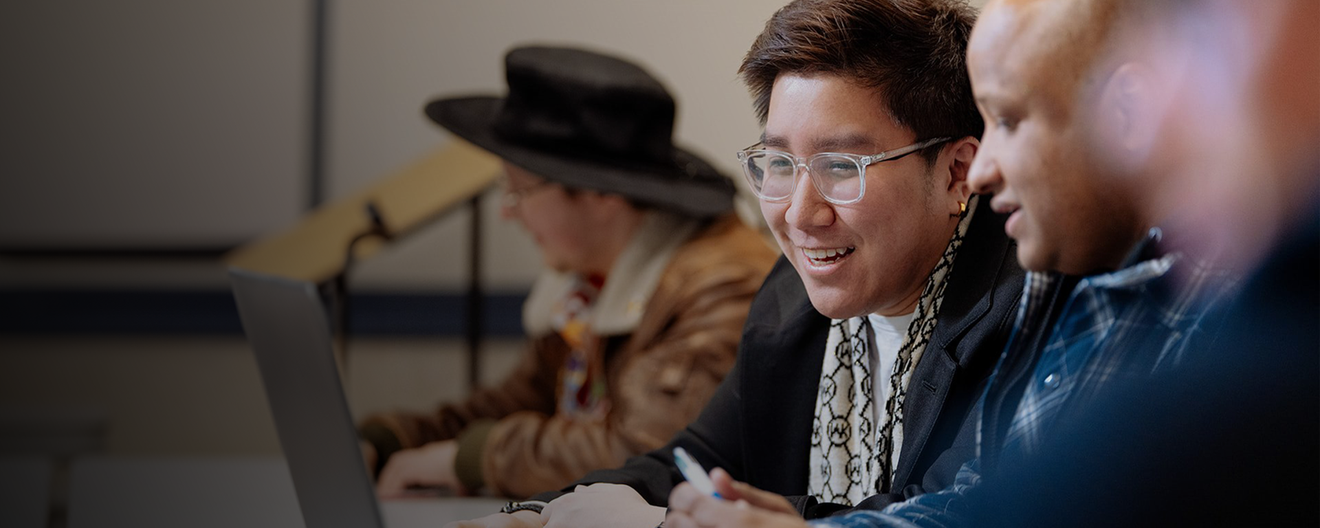 Students are engaged with each other's work in a classroom. Several of them are looking at the same laptop.