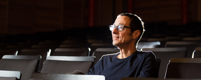 School of Creative and Performing Arts Director Bruce Barton sits in the seats of a darkened theater. He looks towards the stage, and his face is lit.