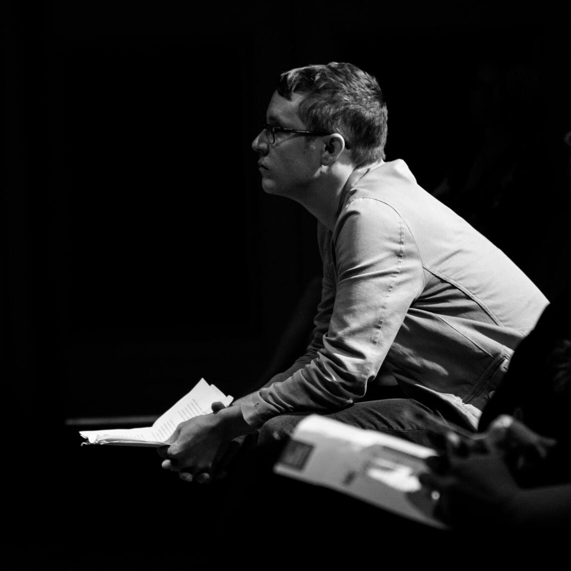 Former Writer-in-Residence Nick Thran in a black and white photo looks thoughtfully to the left, holding a sheet of paper. Photo by Monique de St. Croix.