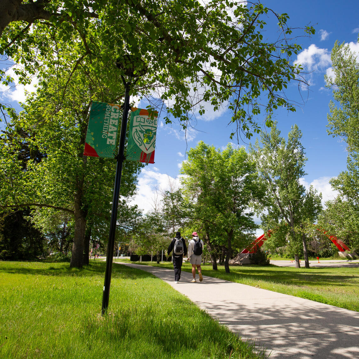 A bright day on campus. Students walk on a pathway. The UCalgary Create Something banners are visible to one side of the path.