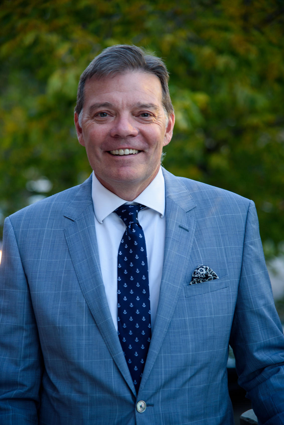 Faculty of Arts Dean Richard Sigurdson poses in a headshot. He is wearing a suit and smiling at the camera. Behind him is green foliage.