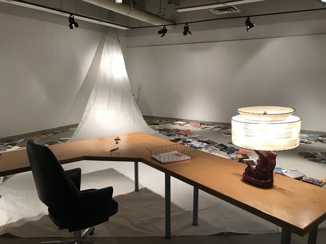 Photo of Brian Rusted exhibition in the 621 Gallery. Visible in the picture is a desk, table and lamp. Books are arrayed on the floor beyond the table, and a sheet is hung to look like a sail.