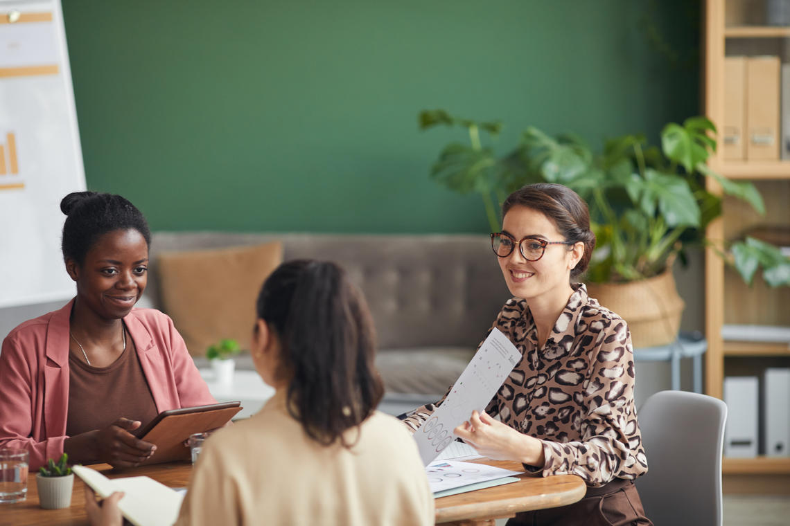 Stock photo of three young women seated around a table in a work setting, engaged in pleasant interaction