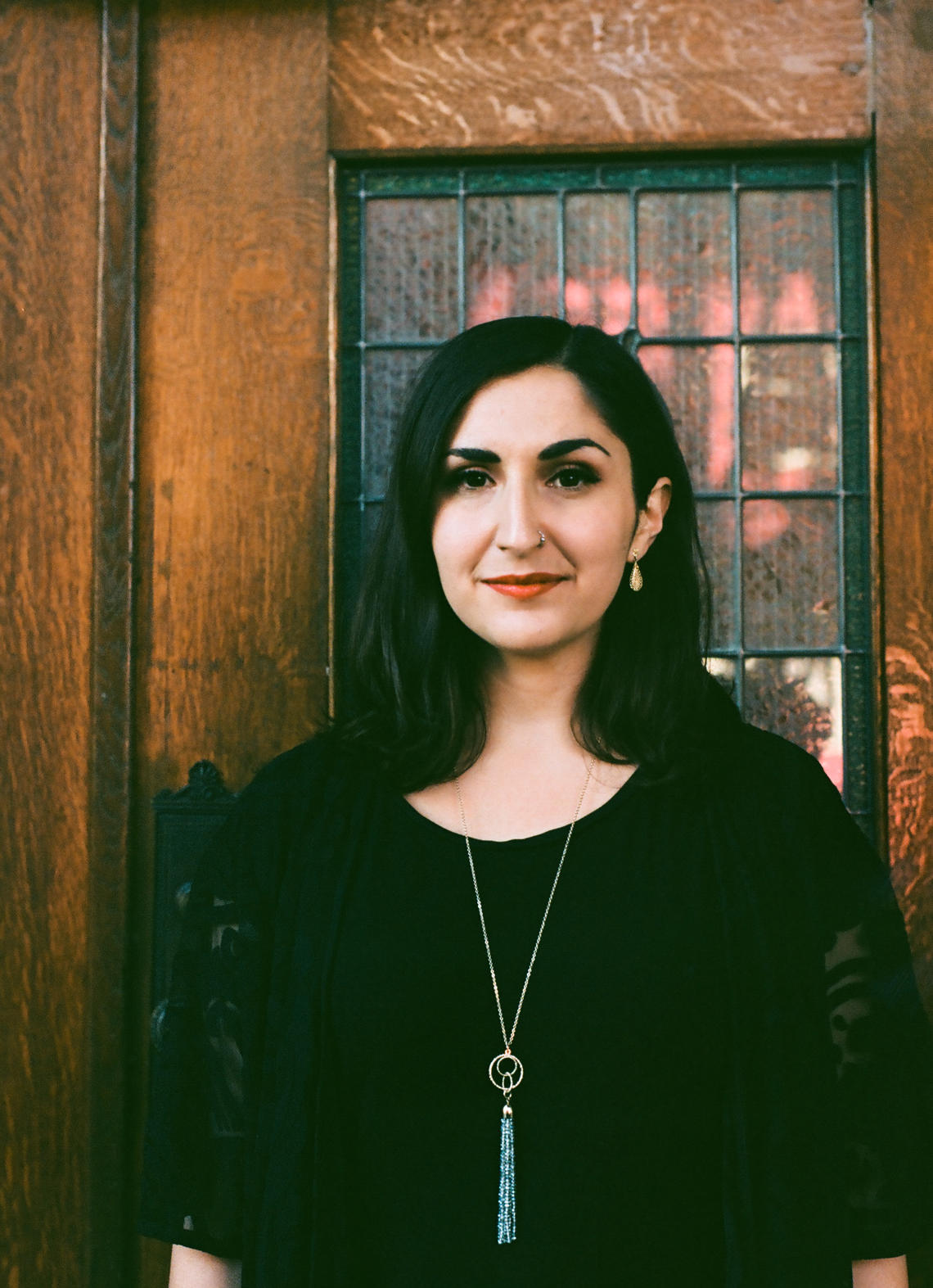 This is a photo of writer Leah Horlick. Leah smiles softly towards the camera with her dark her loose, wearing a black shirt, red lipstick, and gold earrings.