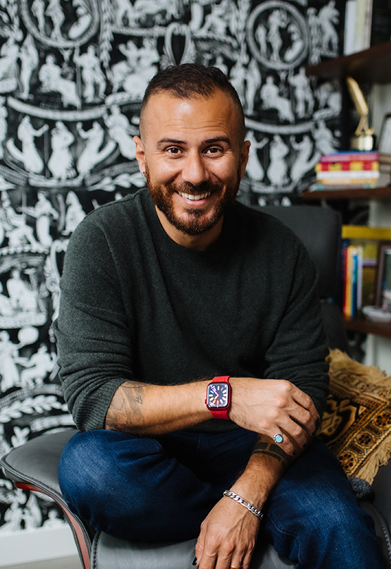 Danny Ramadan sits on a chair, slightly leaning forward and smiling. There is eclectic wallpaper behind him.