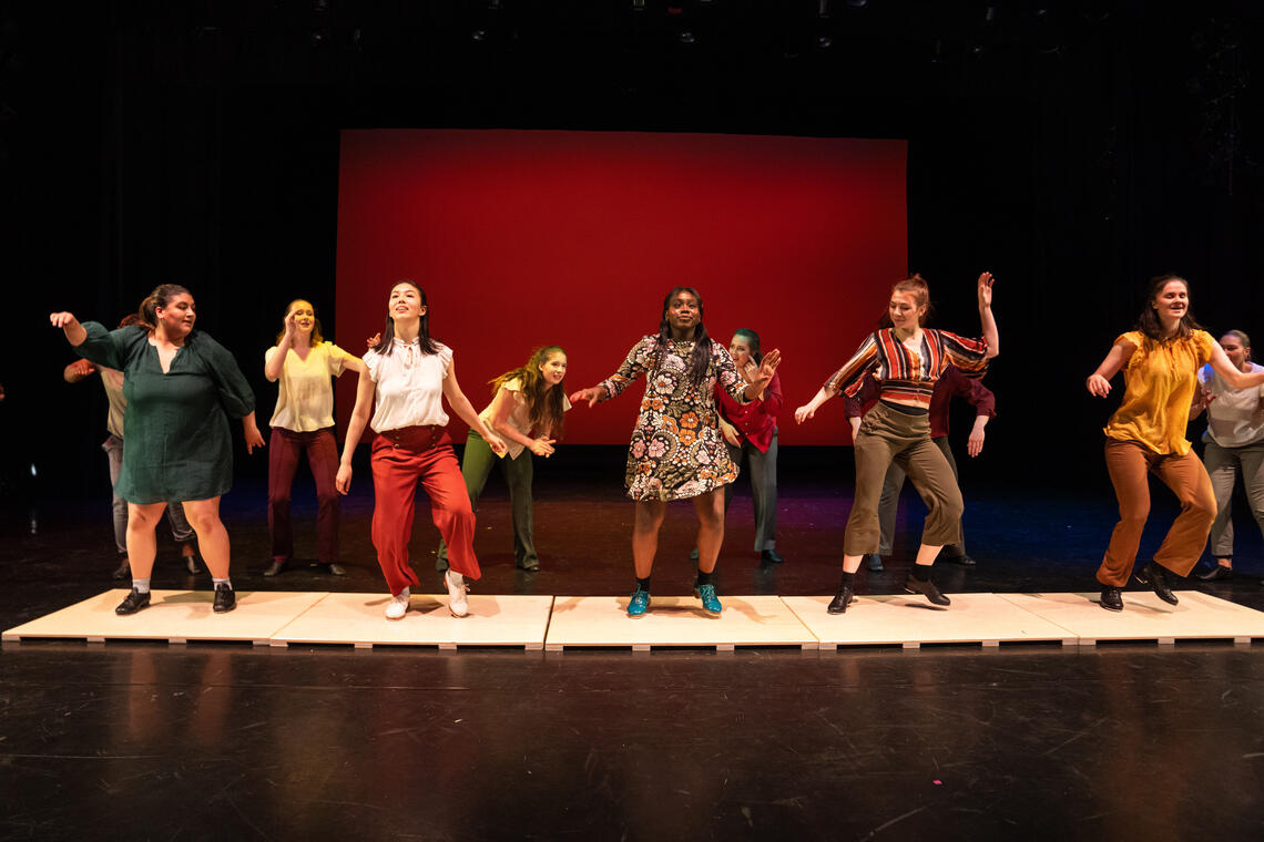 TransFoRM 2025 — there is a row of dancers standing on tap boards facing the audience. The cyc in the background is red.