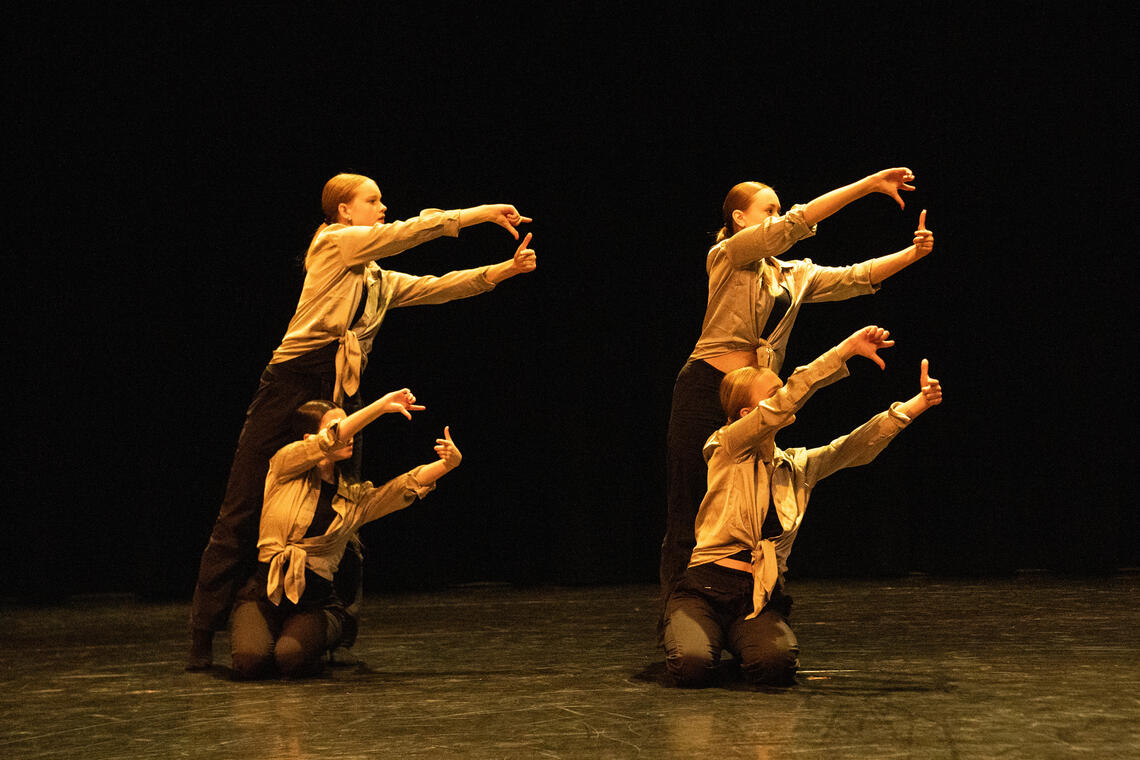 Four dancers, two kneeling and two standing, are reaching to the right with their arms outstretched and hands connecting. They are wearing black pants and brownish button up tops.