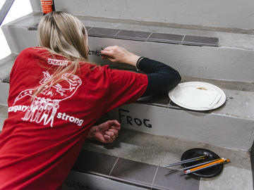 A volunteer wearing a red UCalgary Strong tshirt crouches as she paints a stair