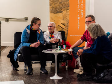 Four people chat around a table, Two of them are queer elders wearing pride scarves.