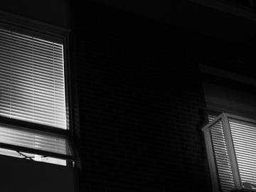 A black and white photo of the outside of a house at night showing two windows with closed venetian blinds. Some light from the inside of the house leaks through the blinds.