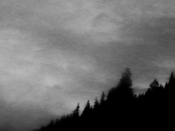 Black and white, grainy photo of a conifer-treed hill at night in the bottom right third of the picture. Clouds cover the rest of the image.