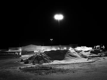 A black and white photo of a Walmart packing lot at night. It is winter and snow has been plowed into a large drift.