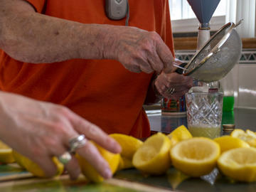 A close up of an elderly woman straining lemon juice into a glass, while a middle-aged woman's hands handle lemons in the unfocused foreground