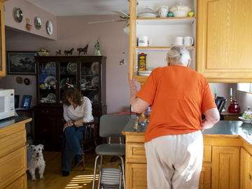 An elderly woman looks in her kitchen cupboards while a middle aged woman sits on a kitchen chair. A dog is beside her.